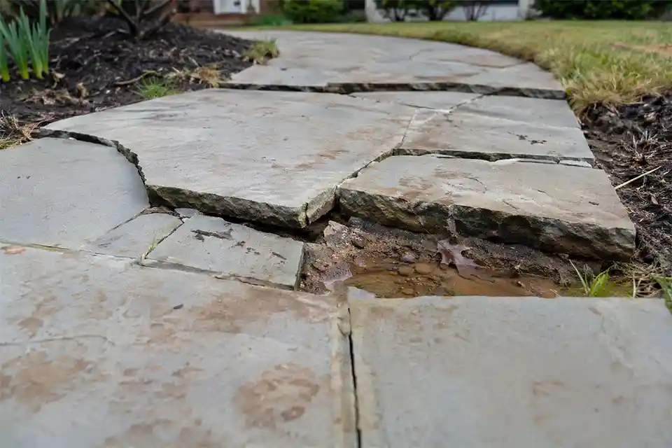 Cracked and slightly sunken outdoor walkway stones breaking over a weak base with widened joints and a shallow low spot